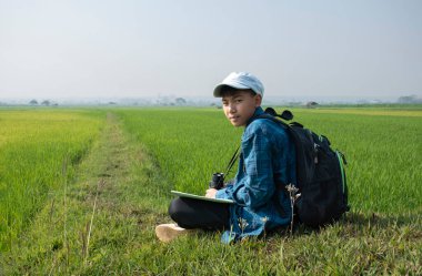 Asian boy wears plaid shirt and cap, has a backpack, binoculars and map, sitting on ridge of rice paddy field to observe the nature near village, birdwatching, pm 2.5 smoke, plants and rice growing.