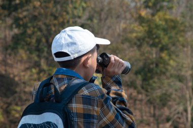Asian boy in plaid shirt wears cap and has a backpack, holding a binoculars, sitting on ridge reservoir in local national park to observe p.m. 2.5 smoke, dust and birds on tree branches and on sky.