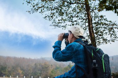 Asian boy in plaid shirt wears cap and has a backpack, holding a binoculars, sitting on ridge reservoir in local national park to observe p.m. 2.5 smoke, dust and birds on tree branches and on sky.