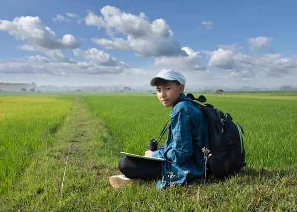 Asian boy wears plaid shirt and cap, has a backpack, binoculars and map, sitting on ridge of rice paddy field to observe the nature near village, birdwatching, pm 2.5 smoke, plants and rice growing.