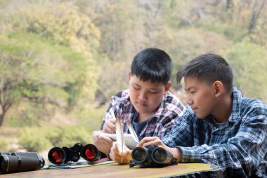 Asian boy in plaid shirt wears cap and has a backpack, holding a binoculars, sitting on ridge reservoir in local national park to observe p.m. 2.5 smoke, dust and birds on tree branches and on sky.