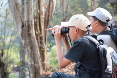 Asian boy in plaid shirt wears cap and has a backpack, holding a binoculars, sitting on ridge reservoir in local national park to observe p.m. 2.5 smoke, dust and birds on tree branches and on sky.