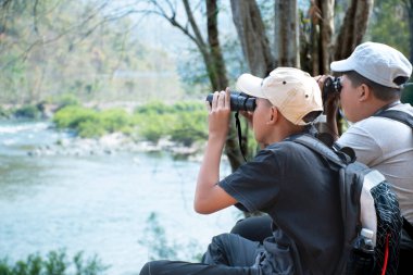 Asian boy in plaid shirt wears cap and has a backpack, holding a binoculars, sitting on ridge reservoir in local national park to observe p.m. 2.5 smoke, dust and birds on tree branches and on sky.