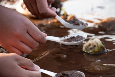 Asyalı çocuk, nehir kıyısındaki çamur, toprak, kaya ve kumları temizlemek için metal kaşık kullanıyor ve içeride yaşayan organizmaları, okul dışındaki bilim laboratuarlarındaki mikropları ve toksinleri inceliyor..