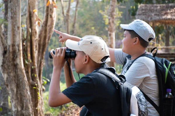 stock image Asian boy in plaid shirt wears cap and has a backpack, holding a binoculars, sitting on ridge reservoir in local national park to observe p.m. 2.5 smoke, dust and birds on tree branches and on sky.