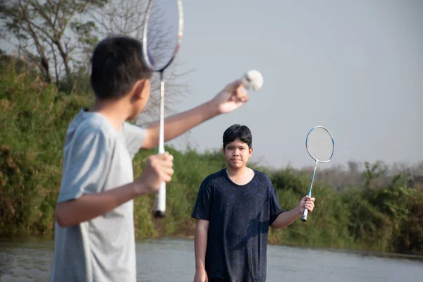 亚洲男孩们在周末放假的时候 拿着羽毛球 羽毛球拍和球拍 站在河边 在他们当地的河边玩耍 他们的注意力是柔软而有选择的 — 图库照片