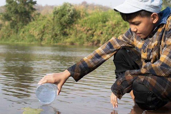 Asian schoolboy holding transparent plastic tube to keep freshwater from local river inside to study the fertility of water, river and fish and to do environment school project work outside the labs.