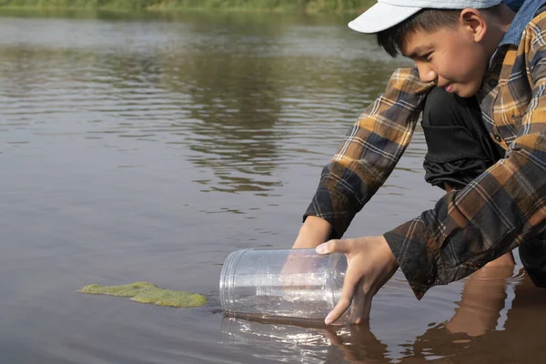 Asian schoolboy holding transparent plastic tube to keep freshwater from local river inside to study the fertility of water, river and fish and to do environment school project work outside the labs.