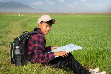 Ekose gömlekli ve kepli Asyalı bir çocuk pirinç tarlasında oturuyor, elinde harita ve dürbün tutuyor, kuşları gözlemlemeden önce harita okuyor, öğleden sonra 2.5 duman ve tarım arazisi sınırları, yumuşak odaklı.