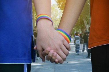 Gökkuşağı bileklikleri takan ve bir grup gencin önünde parkta yürürken el sıkışan Asyalı gay ellerin yakın görüntüsü, LGBT insanları için sevgilerini sunan konsept.