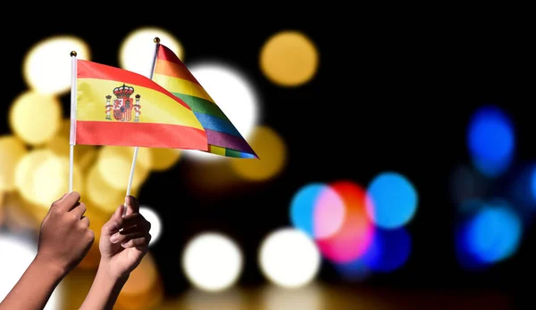 Spain flag and rainbow flag holding in hand with blurred street light bokeh background\', concept for celebrating of LGBT people in Spain in pride month, June.