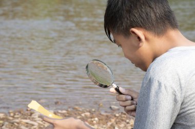 Asyalı çocuk elinde bir büyüteçle oturur ve dışbükey merceğe bakarak öğleden sonra okul tatilinde nehrin kenarında güneş ışınlarından çıkan küçük bir kağıt parçasını deneyler yapmak için yakmaya çalışır..
