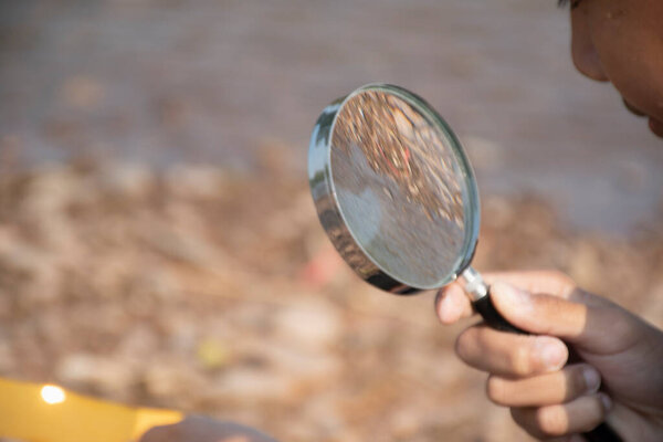 Азиатский мальчик сидит, держа увеличительное стекло и глядя на выпуклые линзы, чтобы попытаться сжечь маленький кусочек бумаги от солнечных лучей возле реки во второй половине дня во время школьных каникул, чтобы сделать эксперимент.