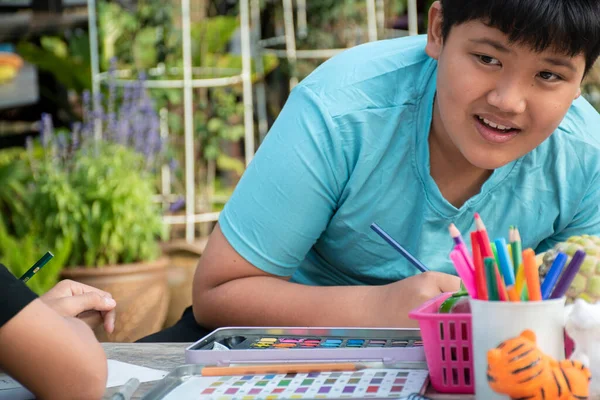 stock image Two boys are spending their free times with drawing, sketching, coloring by using watercolor in the backyard of their house area, teenagers activity in summer concept.