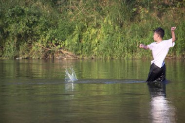 Beyaz tişörtlü Asyalı çocuk boş zamanlarını dalarak, yüzerek, taş atarak ve nehirde mutlu bir şekilde balık tutarak geçiriyor..