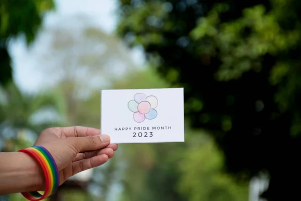 \'Happy Pride Month 2023\' card holding in hand which has rainbow wristband around it, concept for inviting all people to join the LGBTQ+ events around the world in pride month.