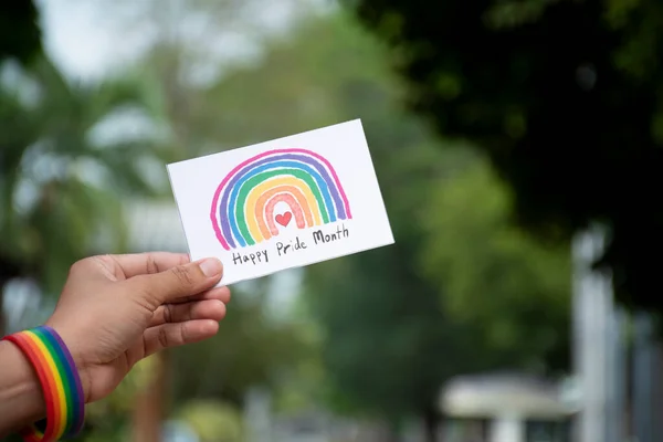 \'Happy Pride Month\' card holding in hand which has rainbow wristband around it, concept for inviting all people to join the LGBTQ+ events around the world in pride month.