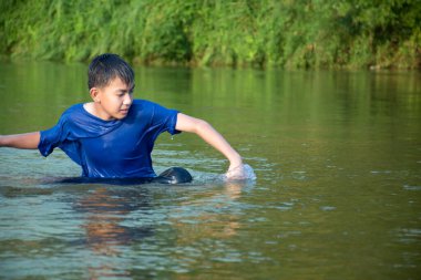 Mavi tişörtlü Asyalı çocuk boş zamanlarını dalarak, yüzerek, taş atarak ve nehirde mutlu bir şekilde balık tutarak geçiriyor..