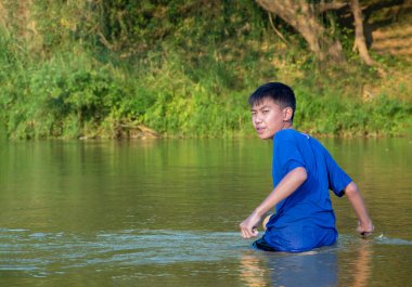 Mavi tişörtlü Asyalı çocuk boş zamanlarını dalarak, yüzerek, taş atarak ve nehirde mutlu bir şekilde balık tutarak geçiriyor..