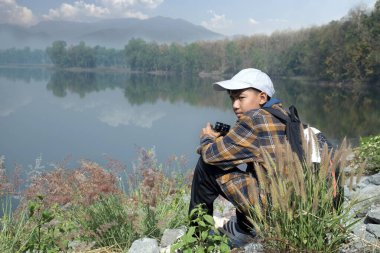 Ekose gömlekli Asyalı çocuk beyaz başlık takıyor, elinde dürbün tutuyor, yaz tatilinde rezervuar tepesinde oturuyor ve kuş izleme etkinliği, yumuşak ve seçici odaklanma, doğa çalışması ve hobi kavramı..