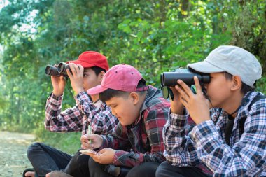 Dürbün tutan Asyalı çocuklar, parkta yaşayan kuşları, böcekleri ve diğer vahşi hayvanları izlemek için yan yana oturuyorlar, solcu ve ortanca çocuklara yumuşak ve seçici bir şekilde odaklanıyorlar, doğa eğitimi kavramı..