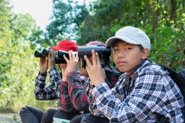 Dürbün tutan Asyalı çocuklar, parkta yaşayan kuşları, böcekleri ve diğer vahşi hayvanları izlemek için yan yana oturuyorlar, solcu ve ortanca çocuklara yumuşak ve seçici bir şekilde odaklanıyorlar, doğa eğitimi kavramı..