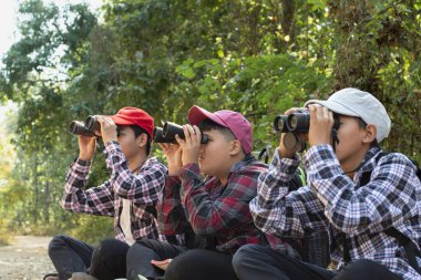 Dürbün tutan Asyalı çocuklar, parkta yaşayan kuşları, böcekleri ve diğer vahşi hayvanları izlemek için yan yana oturuyorlar, solcu ve ortanca çocuklara yumuşak ve seçici bir şekilde odaklanıyorlar, doğa eğitimi kavramı..