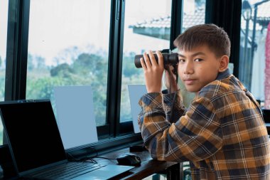 Ekose gömlekli Asyalı yakışıklı çocuk, oturma odasındaki cam pencerenin yanında dürbün tutuyor. Evinin dışındaki ağaçlarda kuşları görmeye hazır. Rahatlama ve eğlence amaçlı etkinlik kavramı..