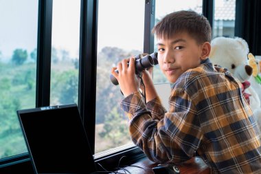 Ekose gömlekli Asyalı yakışıklı çocuk, oturma odasındaki cam pencerenin yanında dürbün tutuyor. Evinin dışındaki ağaçlarda kuşları görmeye hazır. Rahatlama ve eğlence amaçlı etkinlik kavramı..