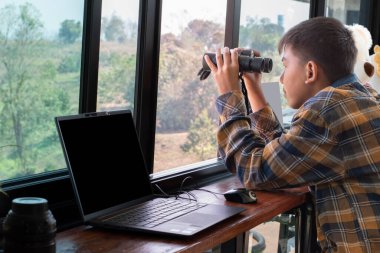 Ekose gömlekli Asyalı yakışıklı çocuk, oturma odasındaki cam pencerenin yanında dürbün tutuyor. Evinin dışındaki ağaçlarda kuşları görmeye hazır. Rahatlama ve eğlence amaçlı etkinlik kavramı..