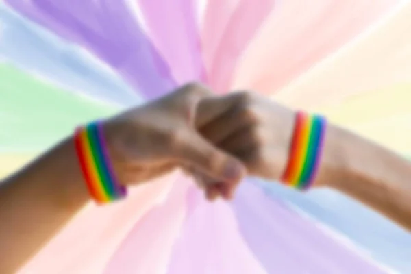 人間の同性への愛とプライド月間イベントのお祝いを示すためのコンセプトである ぼやけたLgbtq フラグの背景を持つ虹のリストバンドを身に着けているゲイカップルのクローズアップ手 — ストック写真