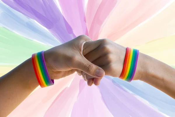 人間の同性への愛とプライド月間イベントのお祝いを示すためのコンセプトである ぼやけたLgbtq フラグの背景を持つ虹のリストバンドを身に着けているゲイカップルのクローズアップ手 — ストック写真