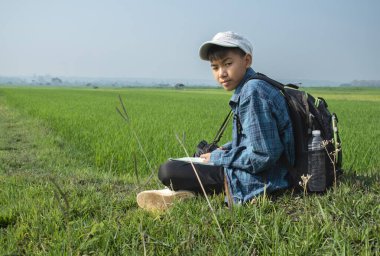 Ekose gömlekli ve şapkalı Asyalı bir çocuk sabahleyin çeltik tarlasında oturuyor ve elinde dürbünle gökyüzünde uçan kuşları izliyor ve pirinç tarlasında balık yiyor, yaz tatili..