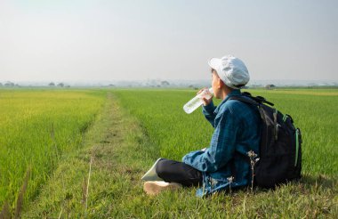 Ekose gömlekli ve kepli Asyalı bir çocuk sabahleyin çeltik tarlasında oturur ve şişeden su içerken gökyüzünde kuşları izler ve pirinç tarlasında balık yer. Yaz tatili..
