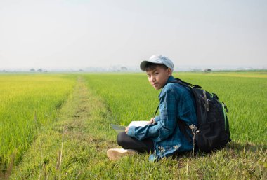 Ekose gömlekli ve şapkalı Asyalı bir çocuk sabahleyin çeltik tarlasında oturuyor ve elinde dürbünle gökyüzünde uçan kuşları izliyor ve pirinç tarlasında balık yiyor, yaz tatili..