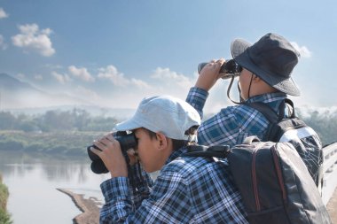 İki Asyalı çocuk, ellerinde dürbünle köprüde duruyor ve aşağıdaki nehirde balıkları ve gökyüzündeki kuşları izliyorlar. Çocuk konseptinin yaz tatili aktiviteleri..