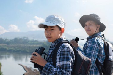İki Asyalı çocuk, ellerinde dürbünle köprüde duruyor ve aşağıdaki nehirde balıkları ve gökyüzündeki kuşları izliyorlar. Çocuk konseptinin yaz tatili aktiviteleri..