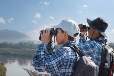 İki Asyalı çocuk, ellerinde dürbünle köprüde duruyor ve aşağıdaki nehirde balıkları ve gökyüzündeki kuşları izliyorlar. Çocuk konseptinin yaz tatili aktiviteleri..