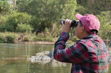 Ekose gömlekli ve kepli Asyalı çocuk yaz etkinliğini kuşlar, balıklar, böcekler, hayvanlar, ağaçlar, çiçekler, harita ve dürbünle izleyerek yapıyor..