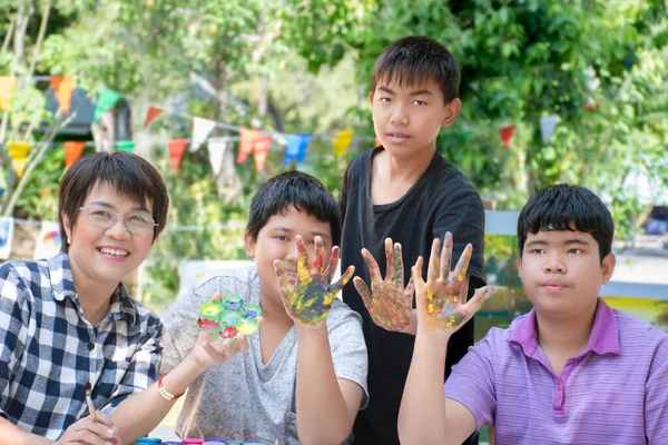 Group of asian boys and female art teacher at outdoor art classrom, summer camp, raising hands which dirty with various colors, smiling happily, concept for summer camps and happy family of people.