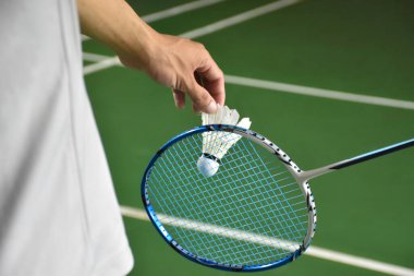 Badminton oyuncusu, raketi ve beyaz kremalı mekikçiyi sahanın diğer tarafına göndermeden önce elinde tutuyor. Tekler konsepte hizmet ediyor..