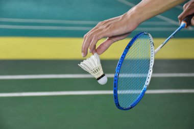Badminton oyuncusu, raketi ve beyaz kremalı mekikçiyi sahanın diğer tarafına göndermeden önce elinde tutuyor. Tekler konsepte hizmet ediyor..