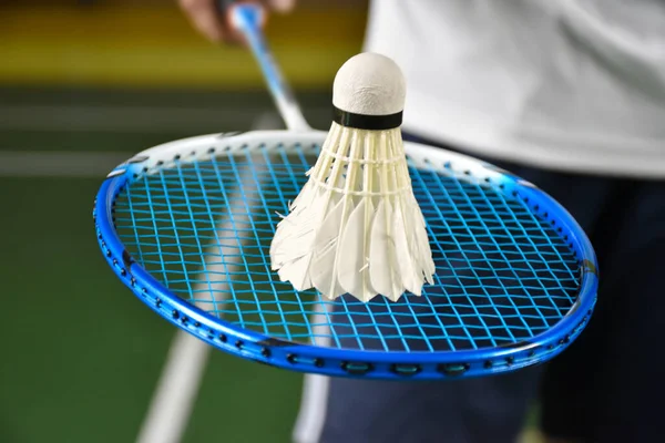 Badminton player receives white cream shuttlecock from service judge by using racket in front of the net before serving it to another side of the court, selective focus on racket and string.