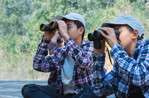 A group of Asian boys sit on floor by the footpath and use binoculars to look at birds flying in the sky and clinging to trees in a national park near their home.