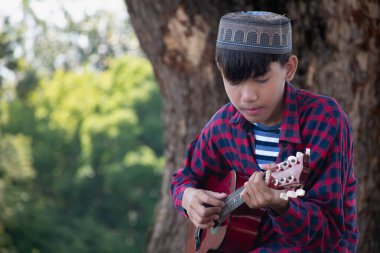 Asyalı tatlı Müslüman ya da İslami çocuk yerel parktaki ağacın altında ukulele çalıyor, eğlence amaçlı aktiviteler yapıyor ve dünyadaki tüm çocukların hobisi.