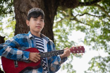 Asyalı tatlı Müslüman ya da İslami çocuk yerel parktaki ağacın altında ukulele çalıyor, eğlence amaçlı aktiviteler yapıyor ve dünyadaki tüm çocukların hobisi.