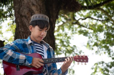 Asyalı tatlı Müslüman ya da İslami çocuk yerel parktaki ağacın altında ukulele çalıyor, eğlence amaçlı aktiviteler yapıyor ve dünyadaki tüm çocukların hobisi.