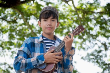 Asyalı tatlı Müslüman ya da İslami çocuk yerel parktaki ağacın altında ukulele çalıyor, eğlence amaçlı aktiviteler yapıyor ve dünyadaki tüm çocukların hobisi.