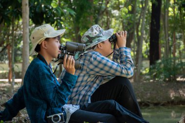 Asian boys are using binoculars to do the birds' watching in tropical forest during summer camp, idea for learning creatures and wildlife animals and insects outside the classroom.