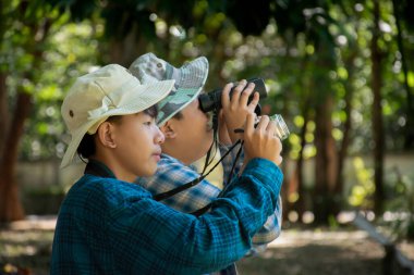 Asian boys are using binoculars to do the birds' watching in tropical forest during summer camp, idea for learning creatures and wildlife animals and insects outside the classroom.
